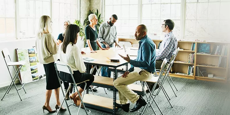 Accelerate Business Innovation : A group of people in a well-lit room engage in a discussion around a table with large windows in the background.