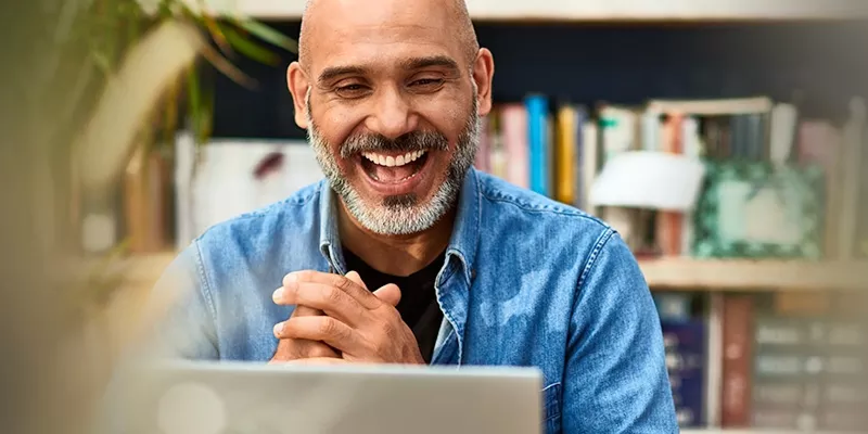 Act on Insights Faster Smiling man in a blue denim shirt in front of a laptop with a bookshelf in the background.